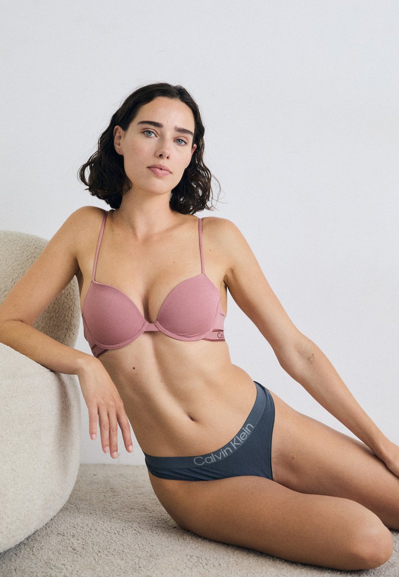En kvinne med kort, bølget hår sitter på en myk, rund sofa og har på seg en rosa BH og grå Calvin Klein undertøy, mens hun ser selvsikkert inn i kameraet.