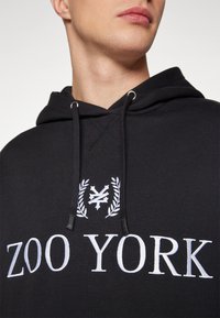 Zwarte capuchontrui met witte tekst en logo "ZOO YORK". Voorzien van aantrekkoordjes en een zachte textuur. Van dichtbij weergegeven, met aandacht voor de details van de halslijn.