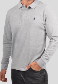 Mies, joka käyttää vaaleanharmaata pitkähihasta polopaidasta, jossa on tumma logo rinnassa ja "USA" kirjailtu hihassa, yhdistettynä mustiin farkkuihin.