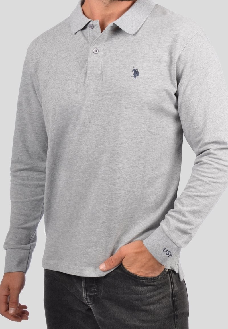 Mies, joka käyttää vaaleanharmaata pitkähihasta polopaidasta, jossa on tumma logo rinnassa ja "USA" kirjailtu hihassa, yhdistettynä mustiin farkkuihin.