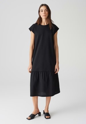 Schwarzes knielanges Kleid mit Kurzärmeln, glattem Oberteil und gerafftem Rüschenshirt. Kombiniert mit schwarzen Absatzsandalen.