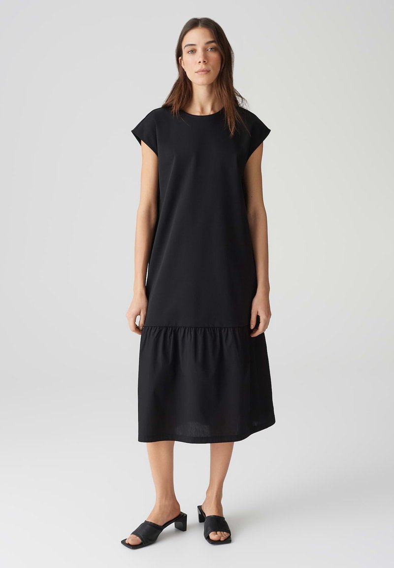 Schwarzes knielanges Kleid mit Kurzärmeln, glattem Oberteil und gerafftem Rüschenshirt. Kombiniert mit schwarzen Absatzsandalen.
