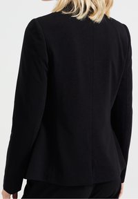 Svart skräddarsydd blazer med en slät textur, strukturerade axlar och en ventilerad baksida. Har långa ärmar och en figurnära silhuett.