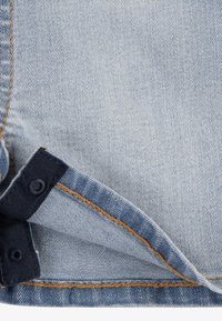 Ljust blå denimtyg med synlig sömnad i orange och mörkblått, med metallknappar i nederkant för justerbar längd.