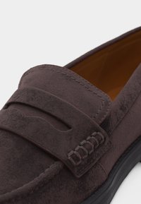 Κοντινό πλάνο ενός σκουρό καφέ μοκασίνι από σουέτ με λουράκι τύπου penny και λεπτομερή ραφές σε μαύρη σόλα.