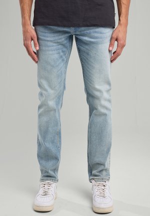 Light blue denim jeans with a slim fit, featuring subtle whiskering and faded areas, paired with white sneakers.
