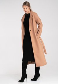 Camelowy płaszcz z wywijanym kołnierzem, pojedynczym guzikiem i bocznymi trokami, noszony na czarnej sukience z prążkowanej dzianiny oraz czarnych kozaczkach do kolan.