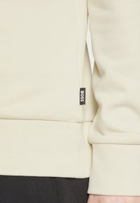 Light beige sweatshirt with textured fabric, ribbed cuffs, and waistband. Features a black "BOSS" tag on the side seam.