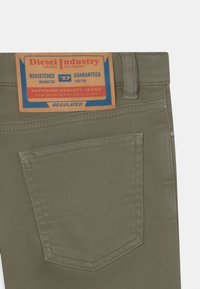 Bolso trasero de unos vaqueros caqui con etiqueta marrón y azul de Diesel Industry que presenta el nombre de la marca, el año de establecimiento 1978 y texto de garantía de calidad.