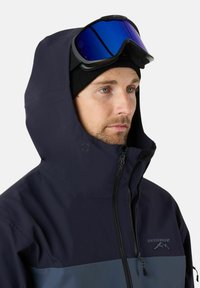 Mężczyzna w czarnej czapce beanie, ciemnoniebieskiej i szarej kurtce z kapturem oraz niebieskich goglach narciarskich podniesionych na czoło, patrzy w prawo.