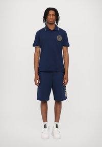 Marineblå polo t-skjorte med gule detaljer og logo, kombinert med marineblå shorts med "FUTURE" i gull. Modellen har på seg hvite joggesko.