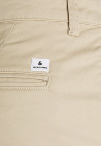 Pantaloni in cotone beige con una texture liscia, dotati di un piccolo tag con il logo JACHS & JONES bianco sulla tasca frontale.