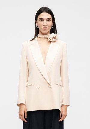 Vrouw met steil donker haar draagt een crème double-breasted blazer en een bijpassende rozenvormige sjaal om haar nek tegen een witte achtergrond.