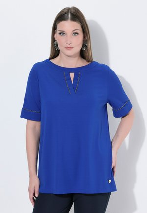 Frau trägt ein königsblaues, kurzärmeliges Oberteil mit V-förmigem Ausschnitt und dekorativem Besatz an den Ärmeln, kombiniert mit schwarzen Hosen.