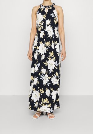 Vrouw draagt een mouwloze zwarte maxi-jurk met een groot wit en beige bloemenpatroon en witte sandalen met blokhak en bandjes.