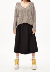 Pull épais en maille de couleur marron clair avec un col en V, associé à une jupe midi noire et fluide. Baskets blanches avec des lacets jaunes et des revers visibles.