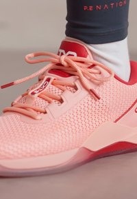 Zapatilla deportiva rosa claro con parte superior de malla, acentos rojos contrastantes, cordones texturizados y una suela gruesa y semitranslúcida.