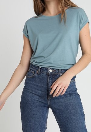 Woman wearing a light blue short-sleeve top tucked into high-waisted dark blue jeans, standing against a plain background.