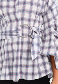 Blusa a quadri bianca, blu e rossa con cintura ad anello argentato e maniche a campana indossata con pantaloni neri.