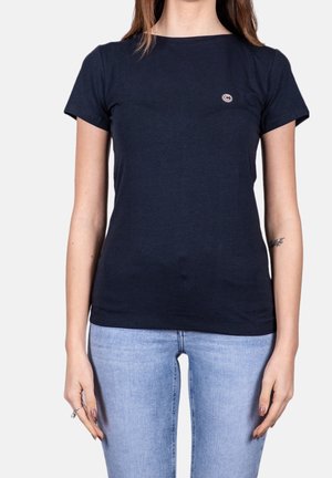 Frau trägt ein schlichtes dunkelblaues T-Shirt mit kleinem Logo auf der Brust und hellblaue Jeans vor hellem Hintergrund.