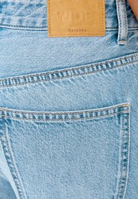 Ljust blå denimjeans med vid passform, försedda med en tan läderlapp märkt "WIDE" och synlig sömnad på bakfickan.