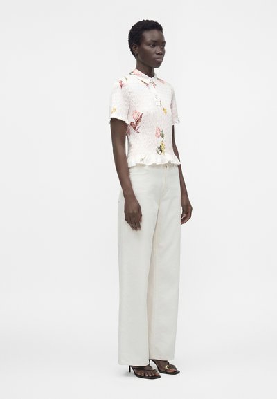 Femme portant un chemisier blanc à manches courtes à motif floral avec un ourlet volanté, un pantalon blanc taille haute à jambes larges, et des sandales noires à talons.
