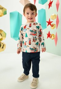 Enfant portant une chemise à manches longues crème avec un imprimé coloré sur le thème des fêtes, associée à un pantalon cargo bleu marine foncé et des baskets en velcro couleur sable.