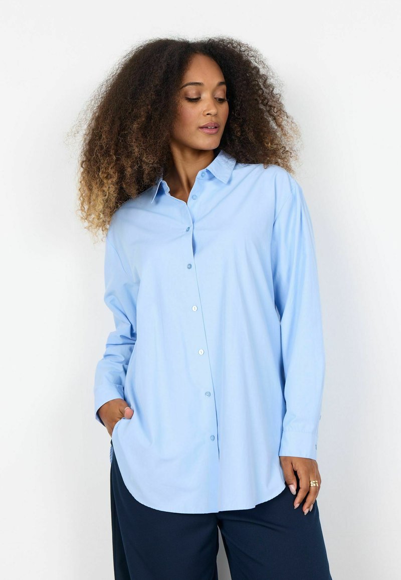 Sieviete ar cirtainiem matiem, valkājot brīvu gaiši zilu pogām aiztaisāmu kreklu un tumšas plataji bikses, stāvot ar vienu roku kabatā.