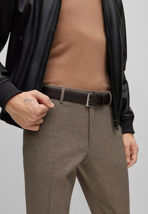 Chaqueta bomber negra, suéter marrón y pantalones marrones con un acabado texturizado. Cinturón de cuero marrón oscuro con una hebilla de metal plateada.