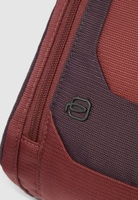 Esquina de bolsa de tela roja y morado oscuro con superficie texturizada, cierre de cremallera y detalle de logotipo en negro. Material duradero con diseño estructurado.