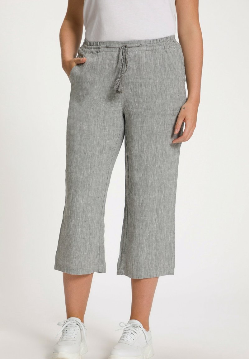 Femme portant un pantalon en lin gris clair coupé au-dessus de la cheville avec taille à cordon, des baskets blanches et un haut sans manches blanc, debout avec les mains détendues.