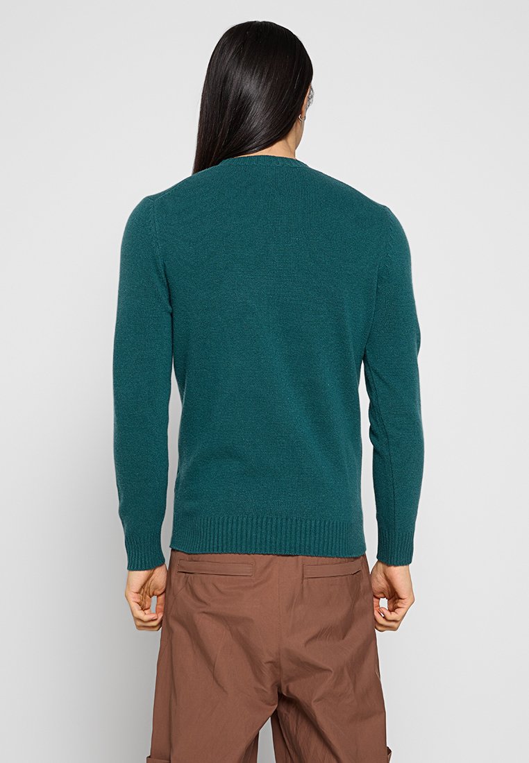 Persoon met lang zwart haar draagt een teal lange mouwen trui en een paarse broek, gezien vanaf de achterkant tegen een effen grijze achtergrond.