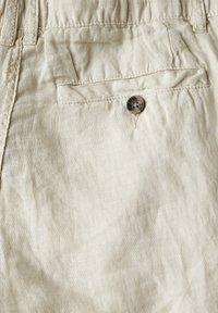 Pantalon en lin beige avec poche arrière à fermeture boutonnée et passants de ceinture visibles.
