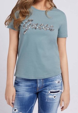 Kvinde iført lyseblå kortærmet t-shirt med dekorative perlebogstaver og slidte blå denimjeans med revner på lårene.