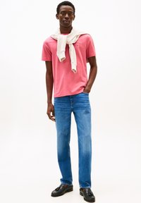 Homme portant un t-shirt rose avec un sweat crème drapé sur les épaules, un jean bleu et des mocassins noirs, se tenant avec une main dans la poche.