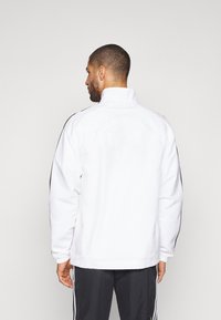 Chaqueta blanca con cuello alto, acentos negros en los hombros y mangas largas. Material suave y ligero. Vista trasera mostrada.