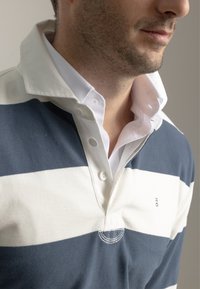 Polo de rayas en azul marino y blanco con cuello abotonado, con una capa inferior blanca. Material de algodón suave con un detalle de logo sutil.