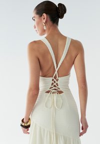 Witte jurk met een gestructureerde stof, diepe V-rug met vetersluiting en brede bandjes. Inclusief gouden armbanden aan de pols.