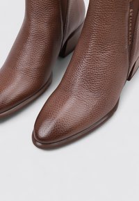 Botas de cuero marrón hasta el tobillo con un acabado texturizado, punteras afiladas y un tacón bajo de apilamiento. Forro interior suave visible en la abertura.