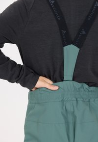 Gröna skidbyxor med elastisk midja och svarta hängslen, som har en slät yta och en lätt design.