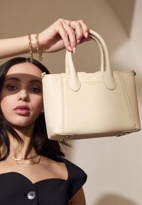 Beige Lederhandtasche mit runden Griffen, geprägtem Branding und goldfarbenen Hardware-Details. In der Hand vor einem neutralen Hintergrund.
