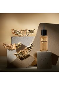 Tapones de perfume con alas doradas exhibidos sobre bloques de piedra junto a una botella recargable de fragancia L'Intense de Zadig & Voltaire.