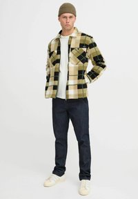 Veste en polaire à carreaux de couleur beige, noire et verte, avec deux poches poitrine et une fermeture éclair à l'avant. Associée à un jean en denim foncé et des baskets blanches.