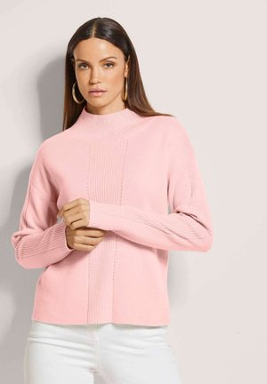 Vrouw met lang bruin haar, draagend gouden oorringen, een roze geribbelde coltrui en witte broek, staand tegen een lichte achtergrond.