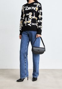 Svart tröja med vita och gula logotyper, kombinerad med blå vidbyxade jeans och svarta stövlar med klack; håller i en liten svart läderhandväska.