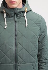 Giacca trapuntata verde chiaro con zip frontale, cappuccio regolabile e dettagli in bianco e beige a contrasto. Texture liscia con tasche.