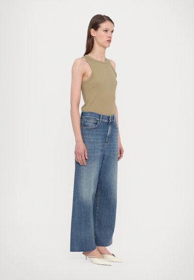 Tank top in olive green fabric, wide-leg blue denim jeans with a faded finish, paired with pointed white shoes.