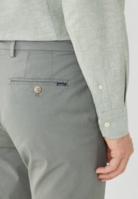 Grijze broek met een knoopzak aan de achterkant en een klein blauw label. Het shirt is lichtgroen met een gestructureerde, geweven uitstraling.
