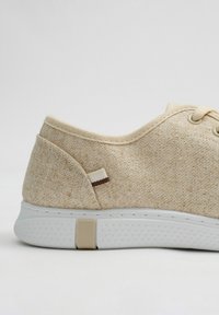 Zapatilla de textil beige con suela de goma blanca, que presenta un pequeño acento marrón y costuras a lo largo del borde. Incluye cordones planos.