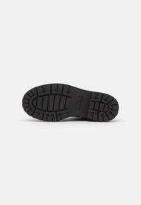 Suela de zapato de goma negra con profundas hendiduras y estrías para tracción, con una forma plana y ancha y un corte rectangular cerca del arco.